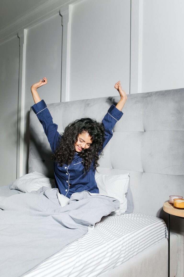 A woman with curly hair enjoying a morning stretch in a cozy bedroom with soft lighting.