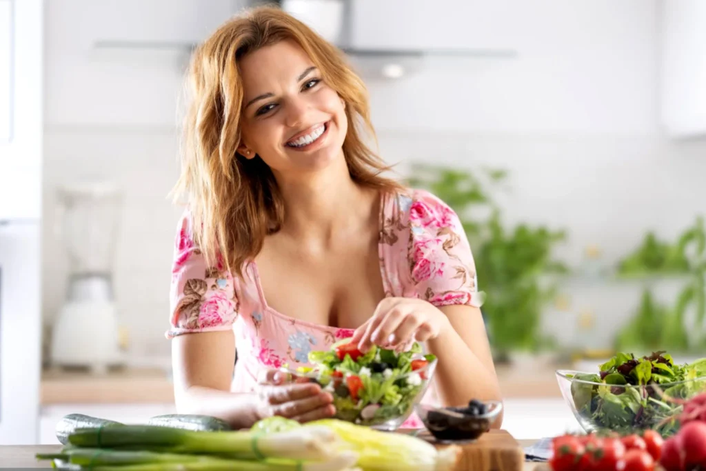 Smiling woman over 40 holding fresh green vegetables and fruits for a healthy flat belly meal plan.